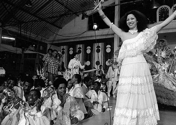Clara Nunes com vestido branco canta em uma quadra de samba.